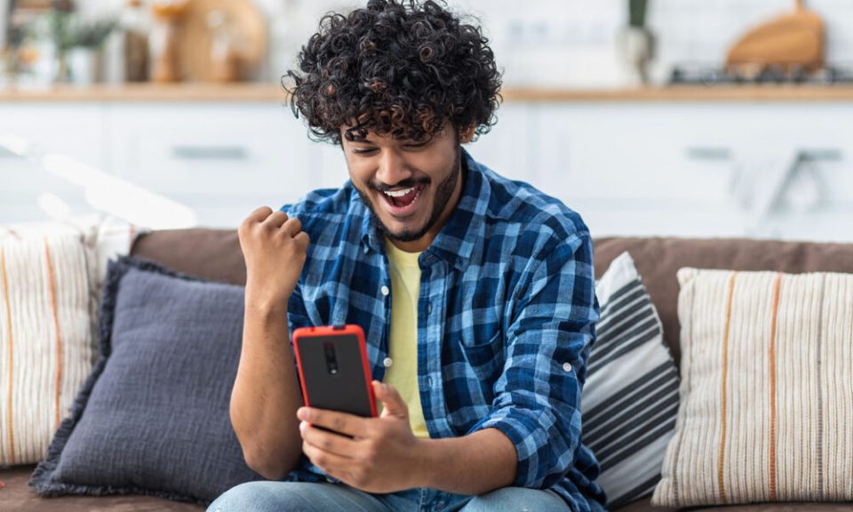Happy Indian male receives online gift feeling joy sitting at home on the couch. Excited young man holding mobile phone reading good news winning lottery game